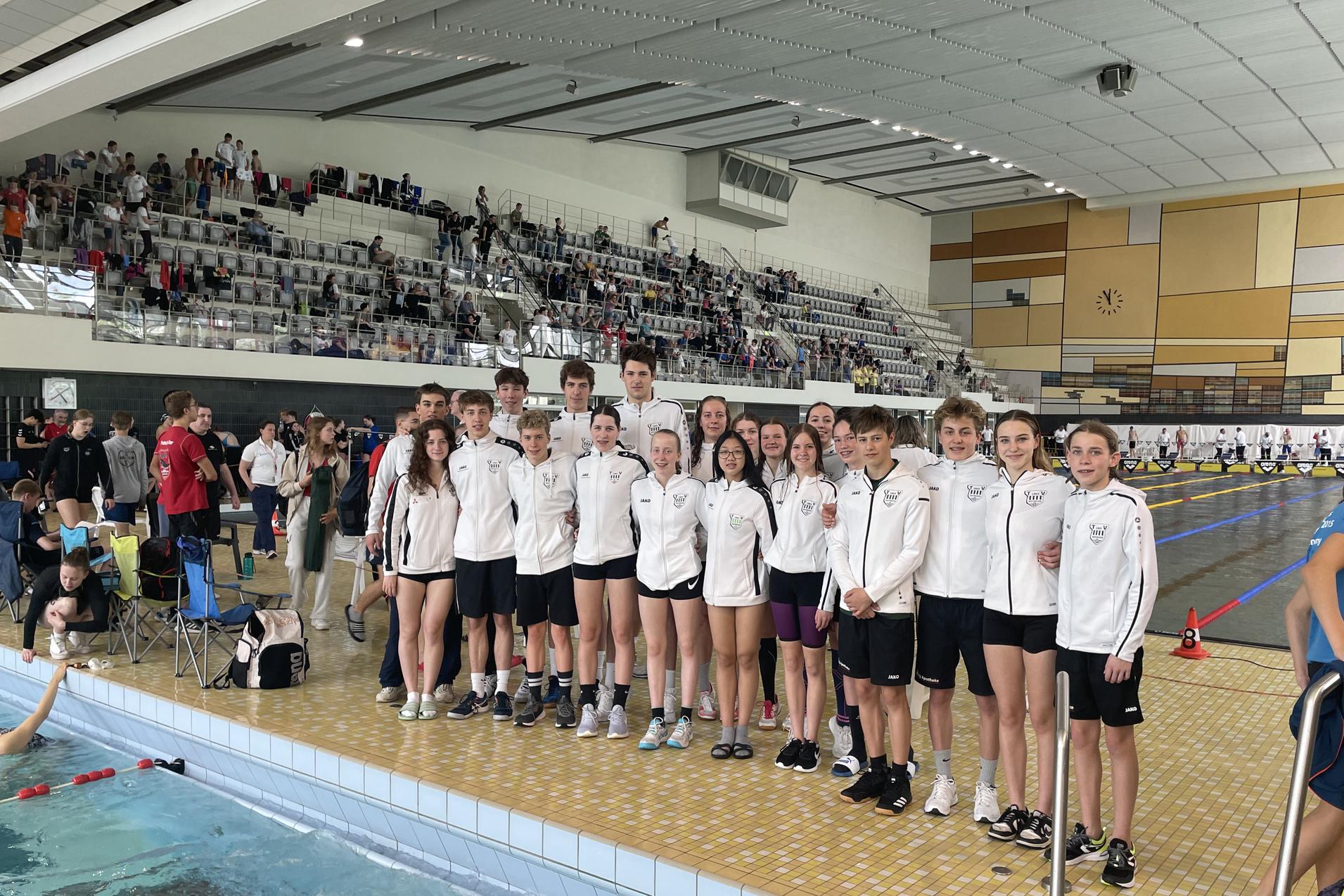 Gruppe von Schwimmern in weißen Jacken steht am Beckenrand in einem Hallenbad mit Zuschauern im Hintergrund.