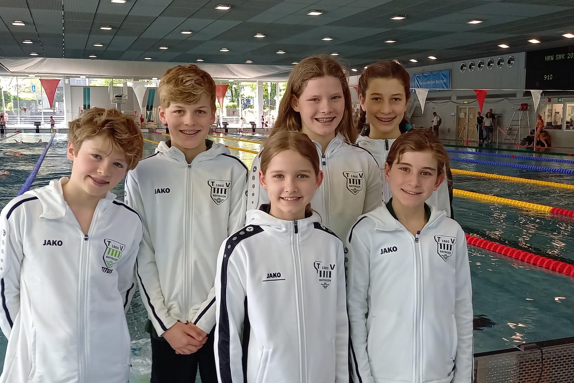 Sechsköpfige Schwimmgruppe in weißen Trainingsjacken am Beckenrand eines Schwimmbads während eines Wettkampfs.