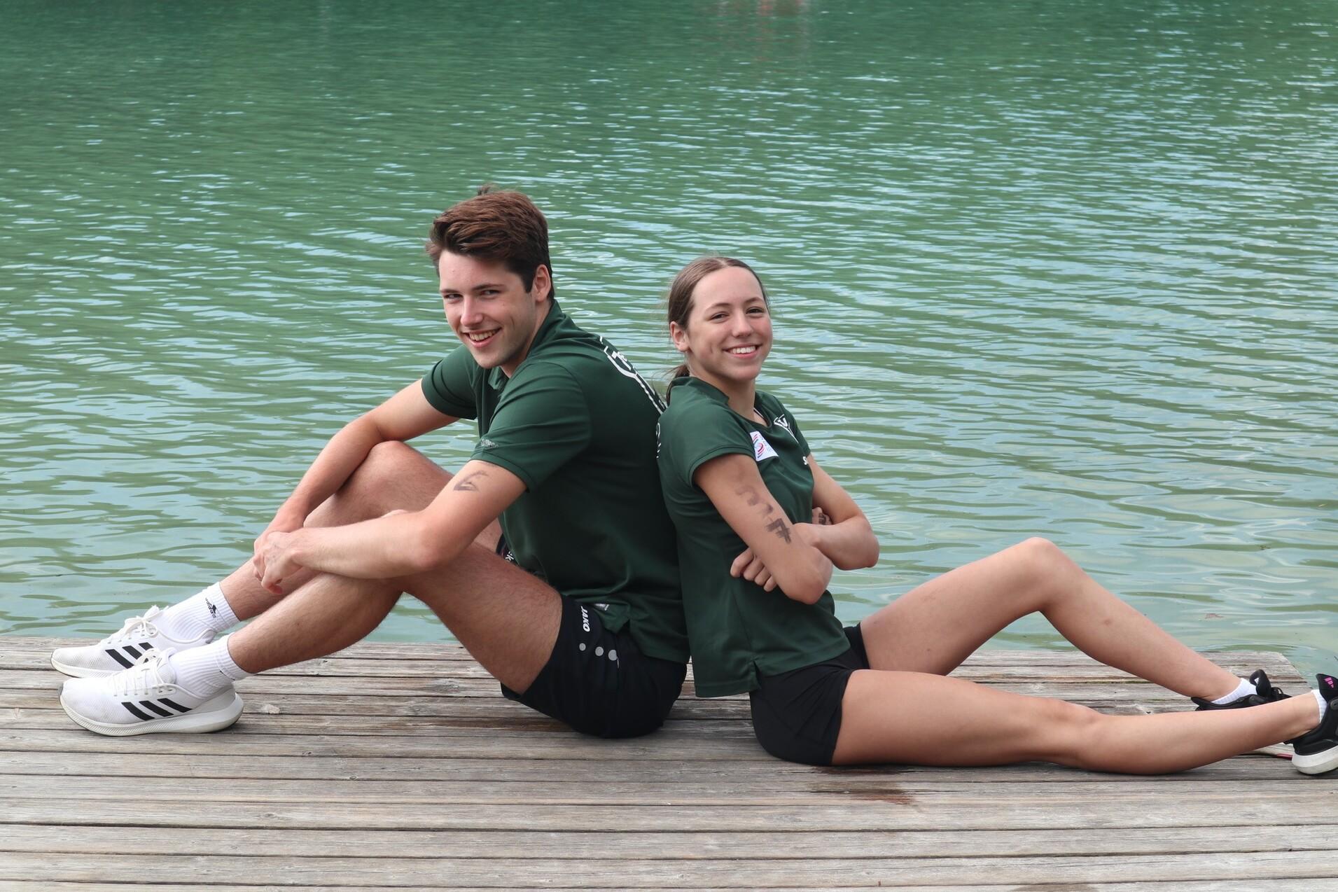 Zwei Sportler sitzen rückwärts an einem Holzsteg mit Wasser im Hintergrund, beide lächeln in die Kamera.