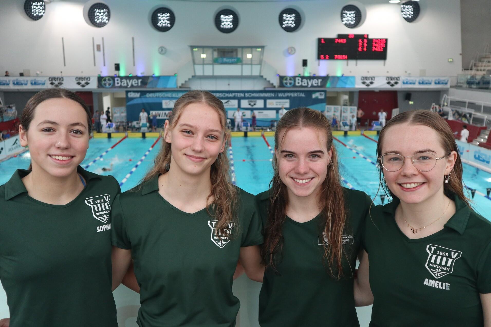 Vier Schwimmerinnen in grünen T-Shirts lächeln vor einer Schwimmhalle mit Wasserbecken und Zeitnahme-Anzeige.