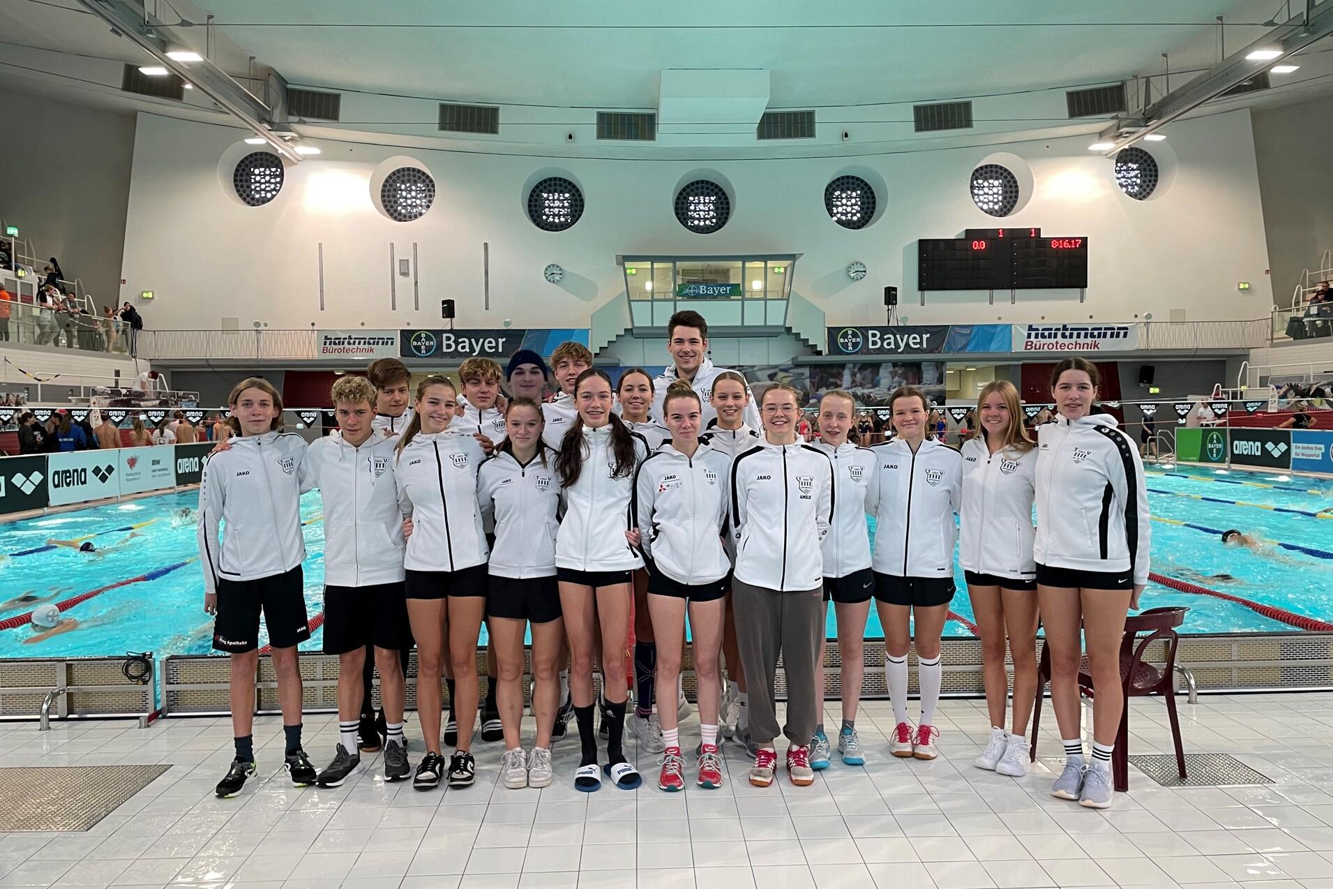 Gruppe von Schwimmern in weißen Jacken, stehend am Beckenrand eines Sportbeckens mit Wettbewerbsatmosphäre.