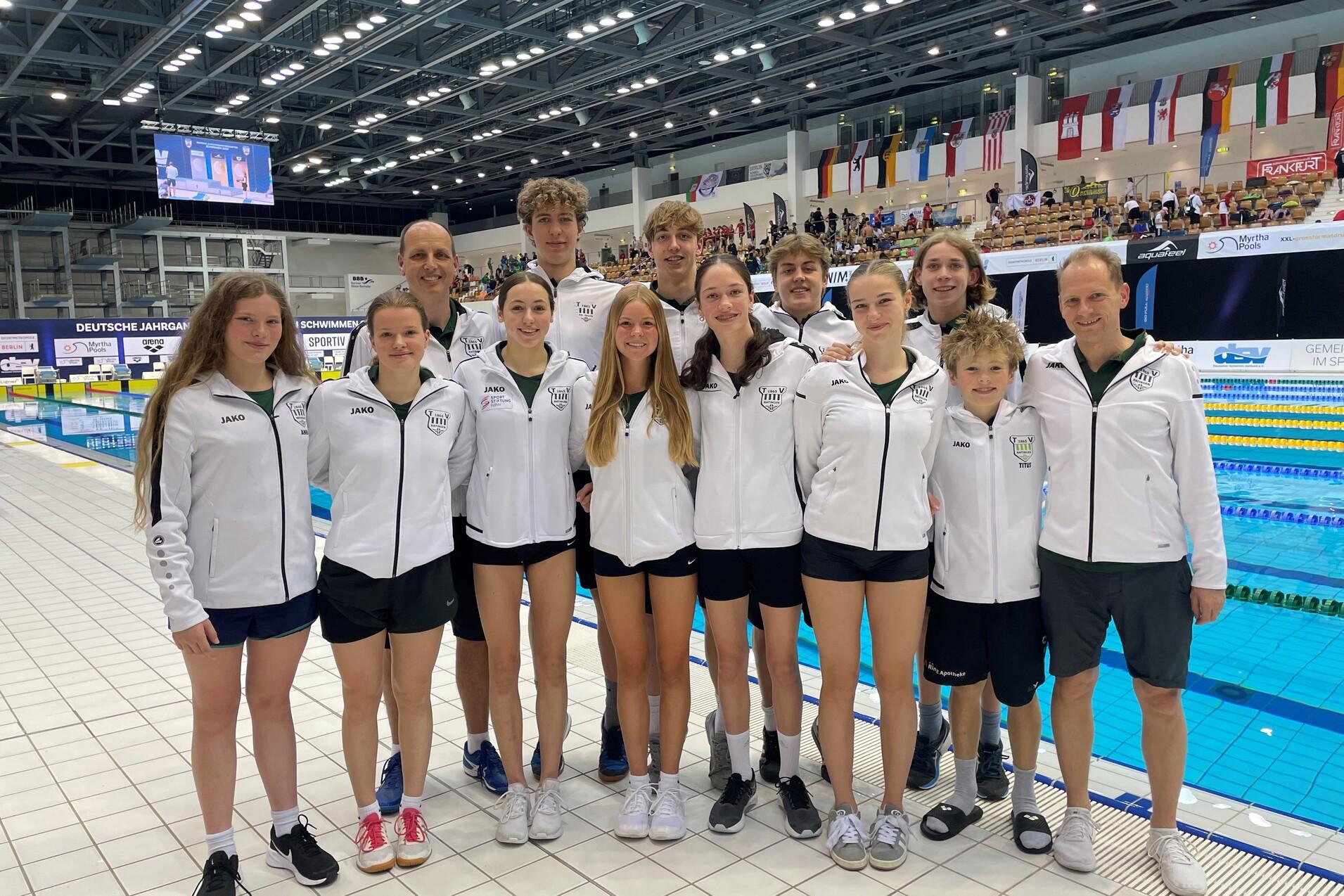 Gruppe junger Schwimmer in weißen Trainingsjacken vor einer Schwimmbeckenauflage bei einem Wettkampf.