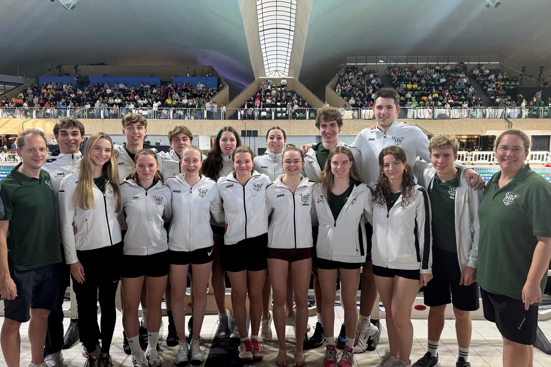 Gruppenfoto von Schwimmern in weißen Jacken und schwarzen Shorts, vor einem Schwimmbecken mit Zuschauern im Hintergrund.