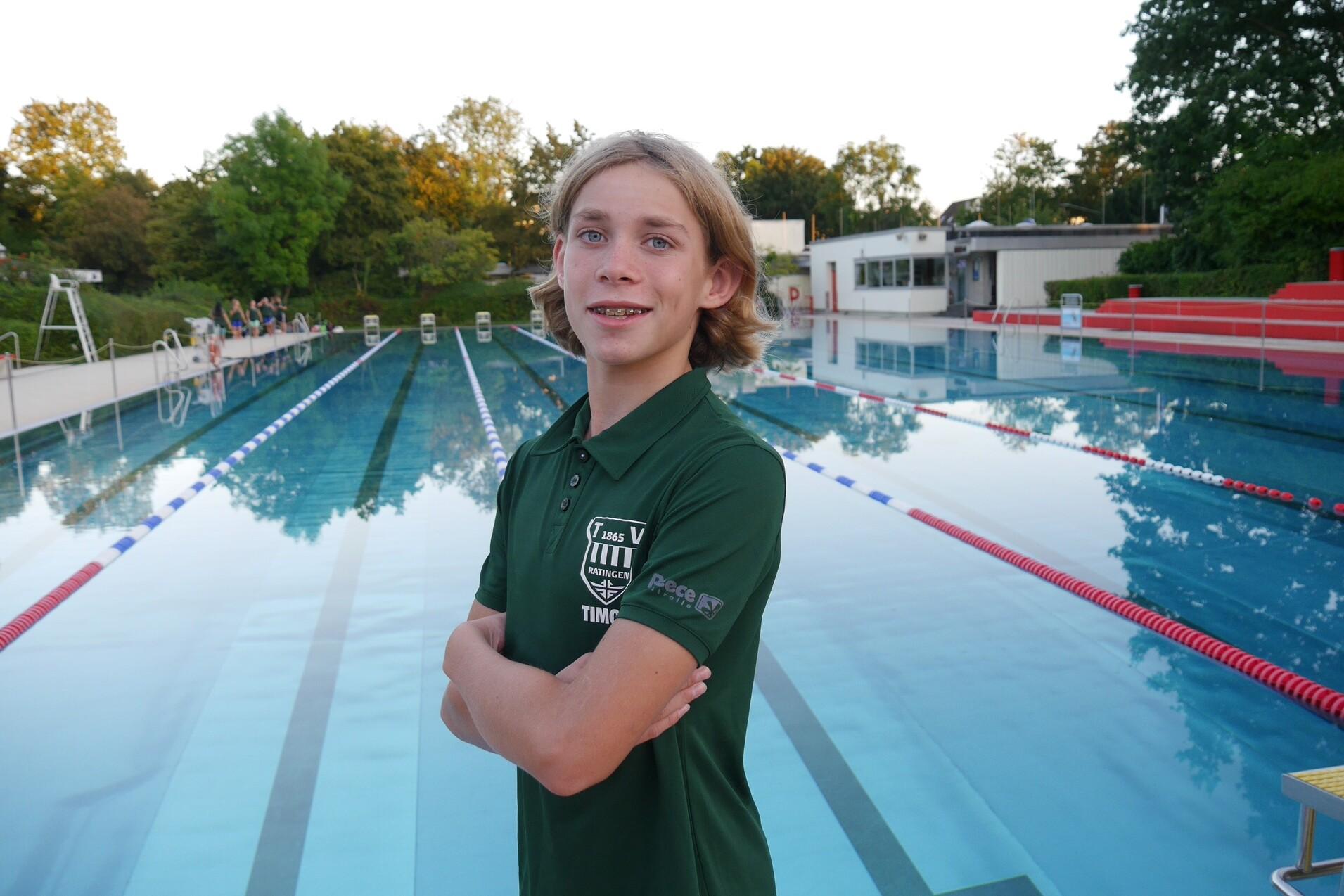 Junger Schwimmer in polo-grünem Shirt steht am Beckenrand eines Schwimmbads mit leeren Bahnen im Hintergrund.