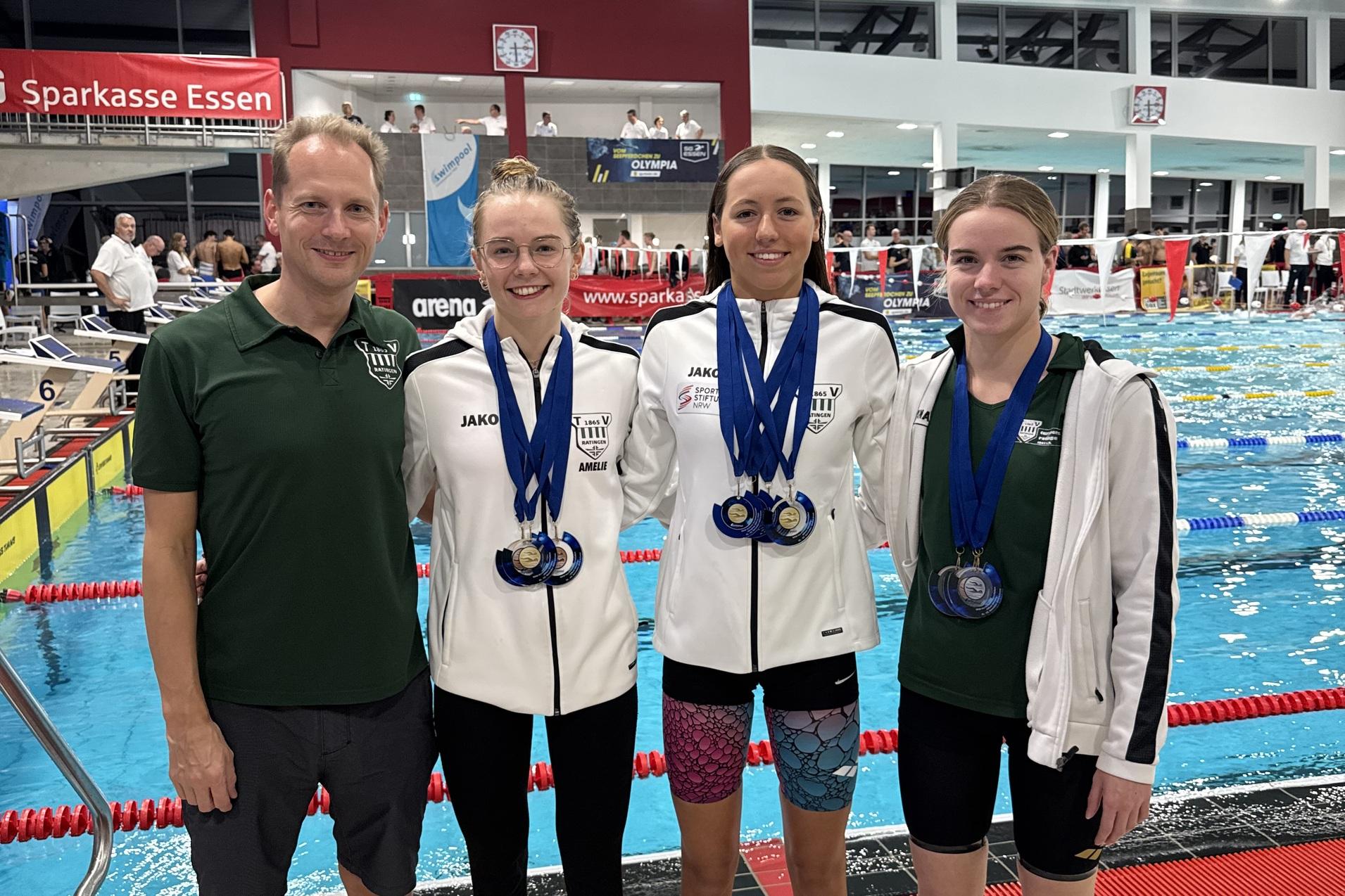 Vier Schwimmerinnen stehen zusammen am Beckenrand, tragen Medaillen und Teamjacken, l&auml;cheln stolz in die Kamera.