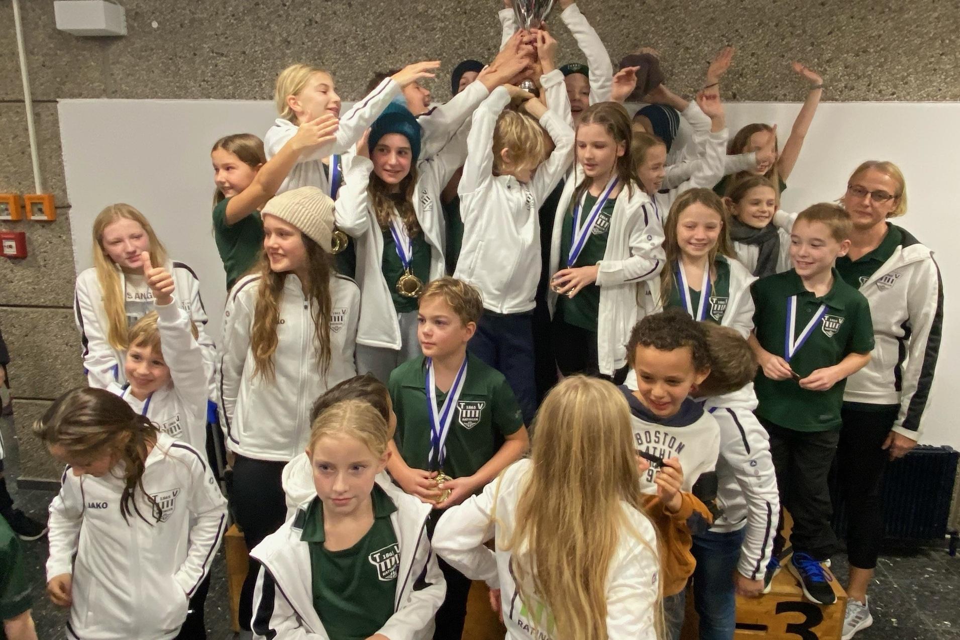 Gruppe von Kindern in Sportjacken feiert mit einem Pokal und Medaillen auf einem Siegertreppchen.