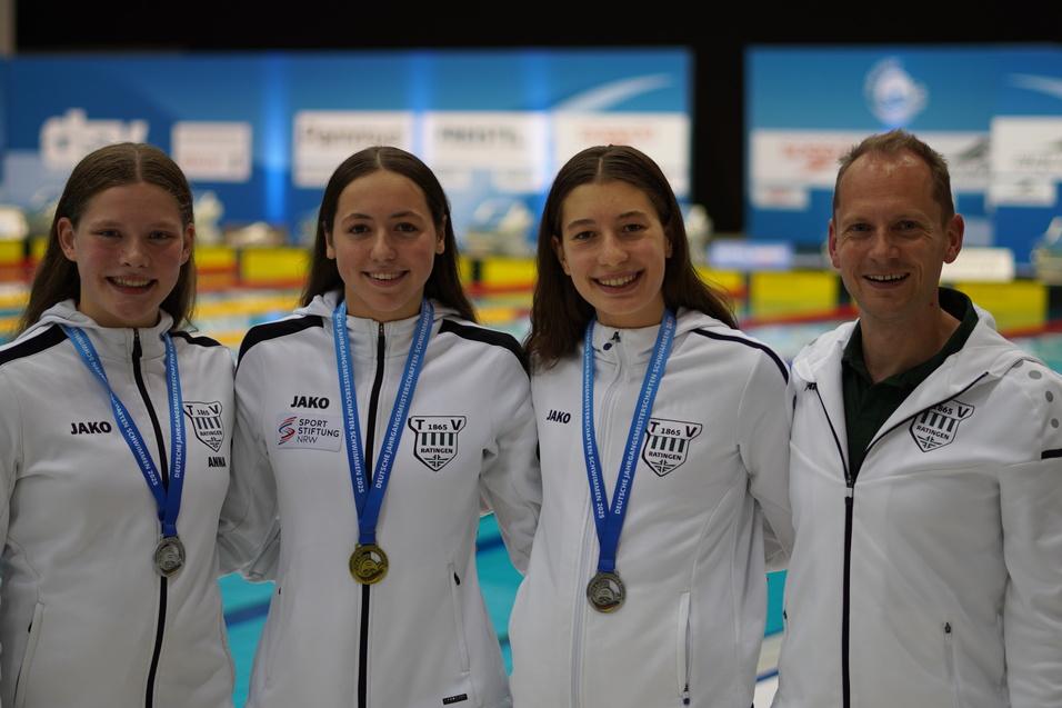 Vier Schwimmerinnen in weißen Trainingsjacken mit Medaillen posieren lächelnd vor einem Schwimmbecken.