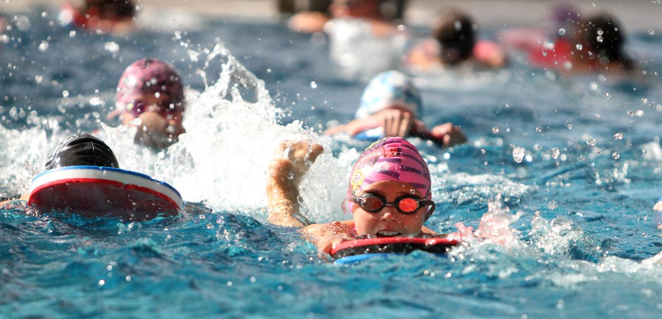 Kinder schwimmen mit Brettern in einem Schwimmbecken, Wasser spritzt auf, einige tragen Schwimmhauben und Brillen.