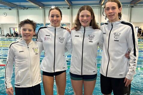 Vier Schwimmer in wei&szlig;en Trainingsjacken stehen am Beckenrand eines Schwimmbads, schwimmend im Hintergrund.