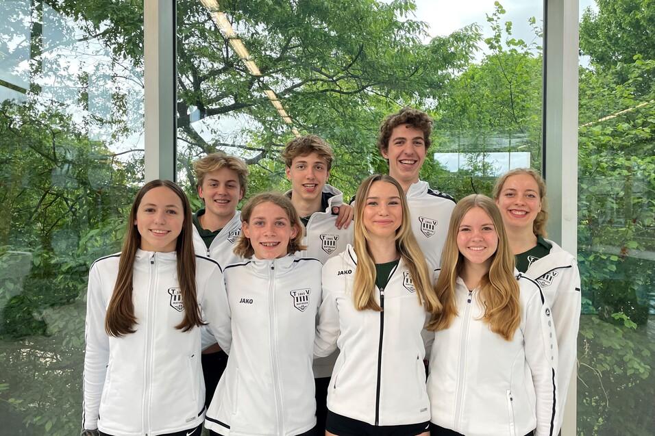Gruppe von acht Jugendlichen in wei&szlig;en Sportjacken mit Mannschaftslogo vor einer gro&szlig;en Fensterfront mit Gr&uuml;npflanzen.