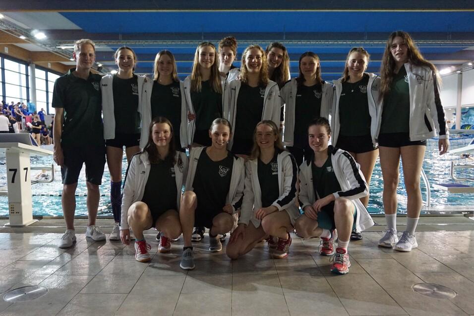 Gruppe von Schwimmerinnen und Trainer am Beckenrand in einer Schwimmhalle, alle tragen Teamjacken und Sportkleidung.