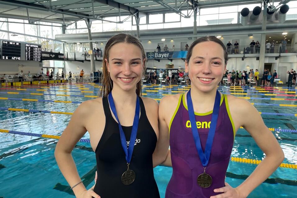 Zwei Schwimmerinnen in Badeanz&uuml;gen mit Medaillen stehen l&auml;chelnd vor einer Schwimmbeckenlandschaft.