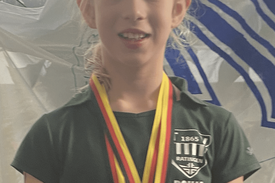 Portr&auml;t eines jungen M&auml;dchens in gr&uuml;ner Sportkleidung mit Medaillen um den Hals vor einem hellen Hintergrund.