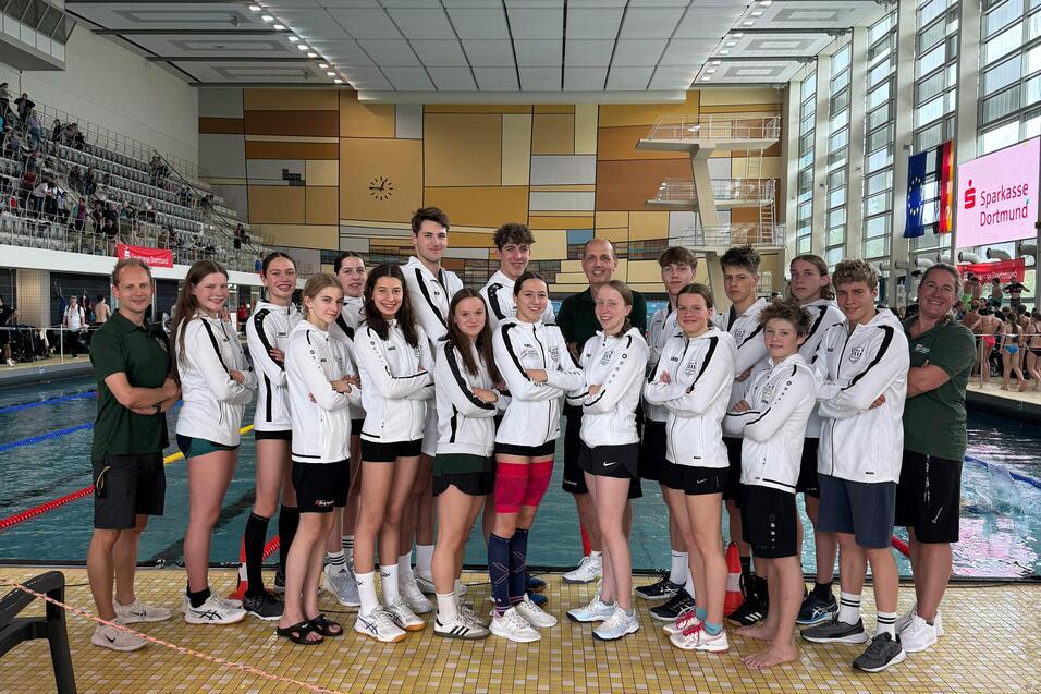 Gruppe von Schwimmern in wei&szlig;en Trainingsjacken posiert am Rand eines Schwimmbeckens in einer Sporthalle.