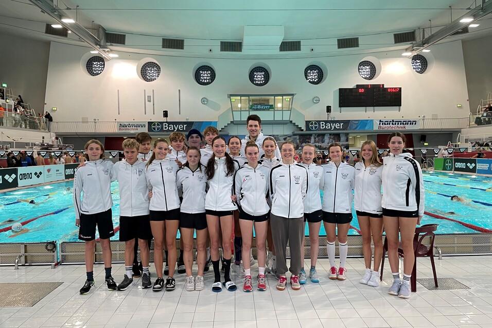 Gruppe von Schwimmern in wei&szlig;en Jacken, stehend am Beckenrand eines Sportbeckens mit Wettbewerbsatmosph&auml;re.
