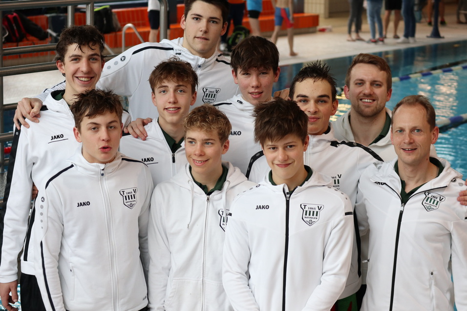 Gruppe von Schwimmern in wei&szlig;en Trainingsjacken, l&auml;chelnd vor einem Schwimmbecken, im Hintergrund weitere Schwimmer.