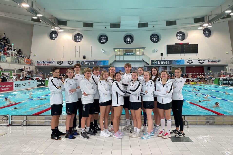 Gruppe von 14 Schwimmern in wei&szlig;en Trainingsjacken vor einem Schwimmbecken mit anderen Schwimmern im Hintergrund.