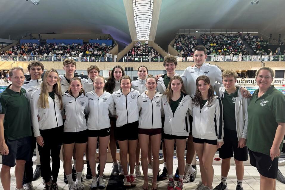 Gruppenfoto von Schwimmern in wei&szlig;en Jacken und schwarzen Shorts, vor einem Schwimmbecken mit Zuschauern im Hintergrund.