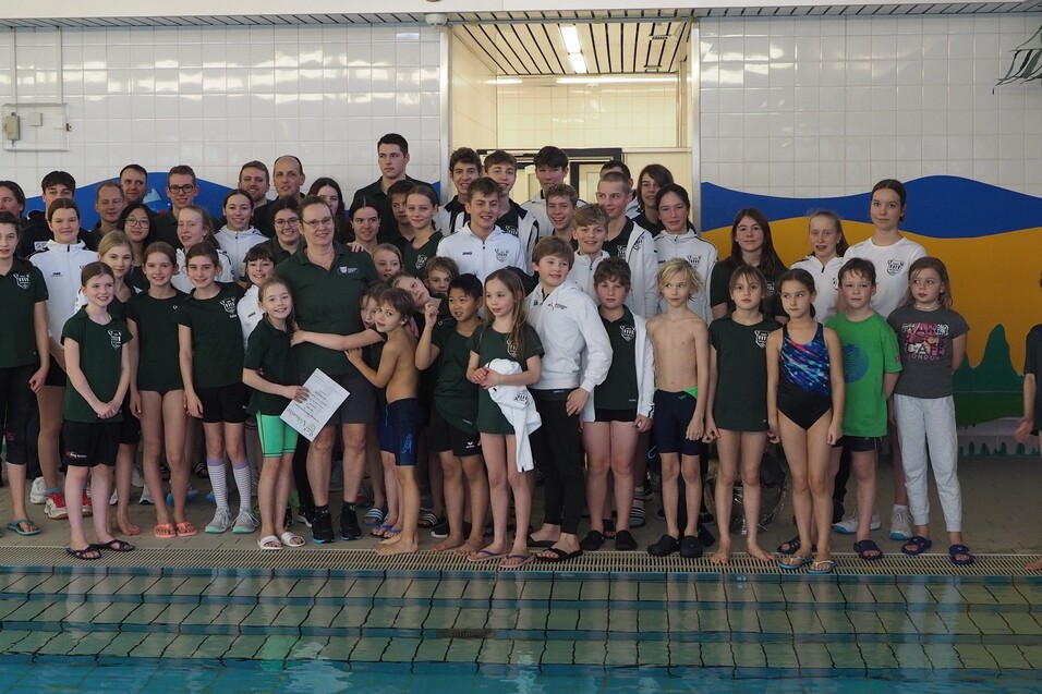 Gruppenfoto von Schwimmern und Trainern in Schwimmbekleidung vor einem Becken mit buntem Wanddesign.