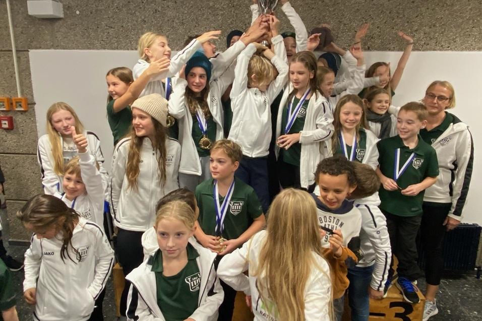 Gruppe von Kindern in Sportjacken feiert mit einem Pokal und Medaillen auf einem Siegertreppchen.