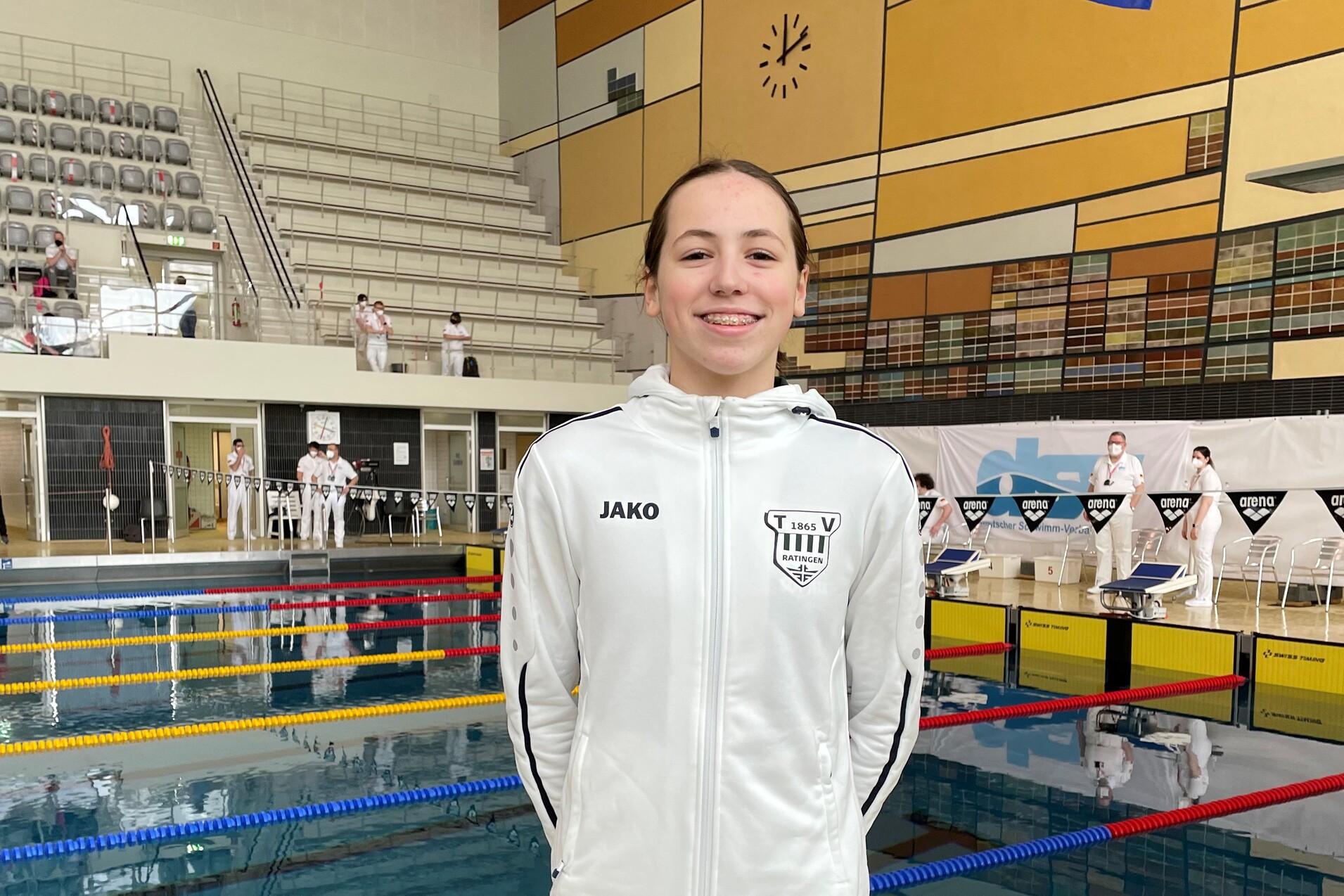 Junge Schwimmerin in weißer Jacke und schwarzer Hose vor einem Schwimmbecken mit Europaflagge im Hintergrund.
