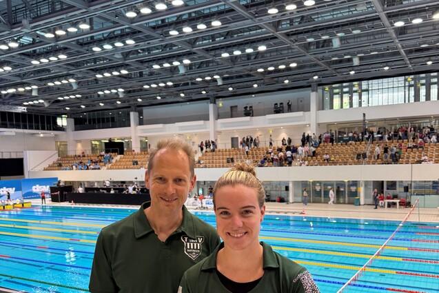 Zwei Personen in gr&uuml;nen Poloshirts stehen vor einem Schwimmbecken mit blauen Wasserbahnen und Zuschauern im Hintergrund.