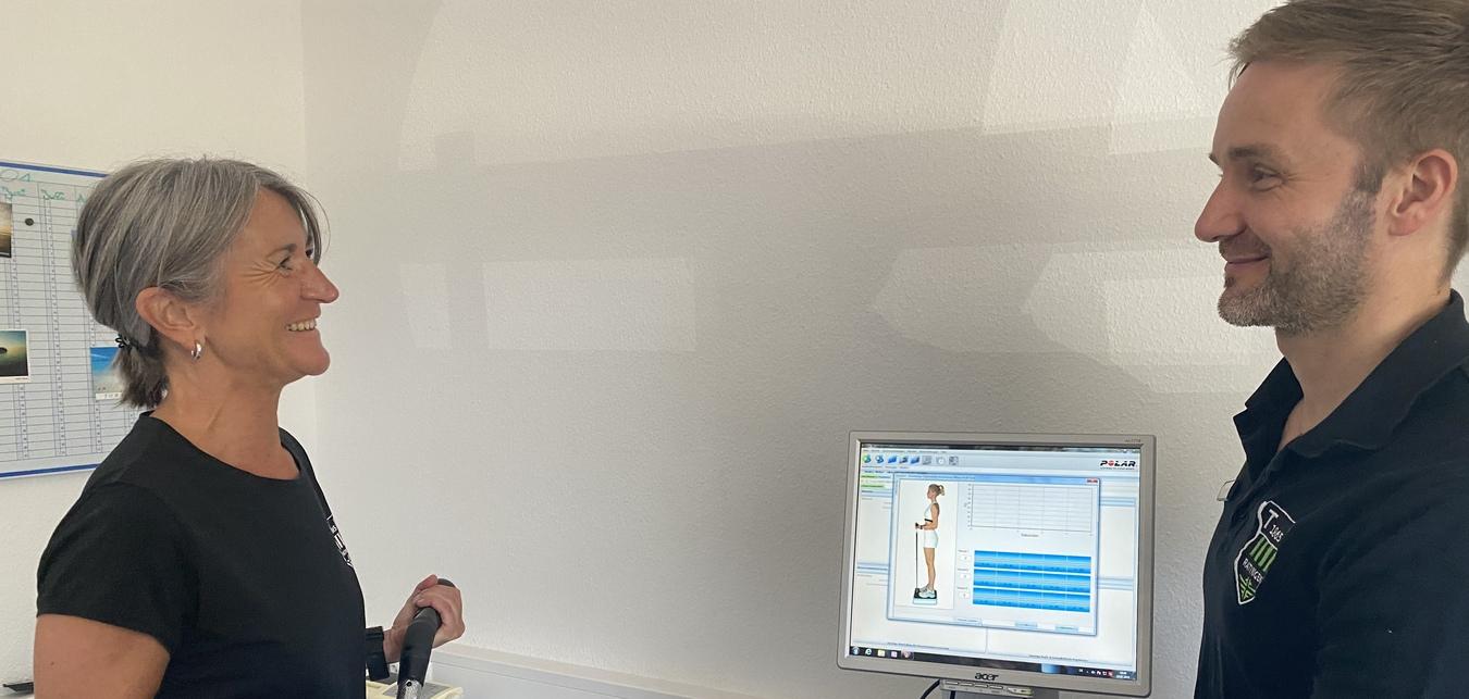 Zwei Personen in einem Untersuchungsraum. Eine Frau h&auml;lt ein Trainingsger&auml;t, w&auml;hrend ein Mann sie an einem Computer ber&auml;t.