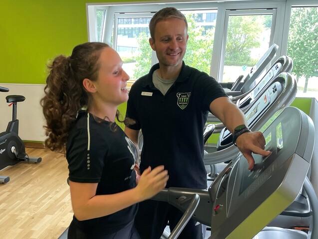 Trainer erkl&auml;rt einer sportlichen Frau die Nutzung eines Laufbandes in einem Fitnessstudio mit gr&uuml;ner Wand.