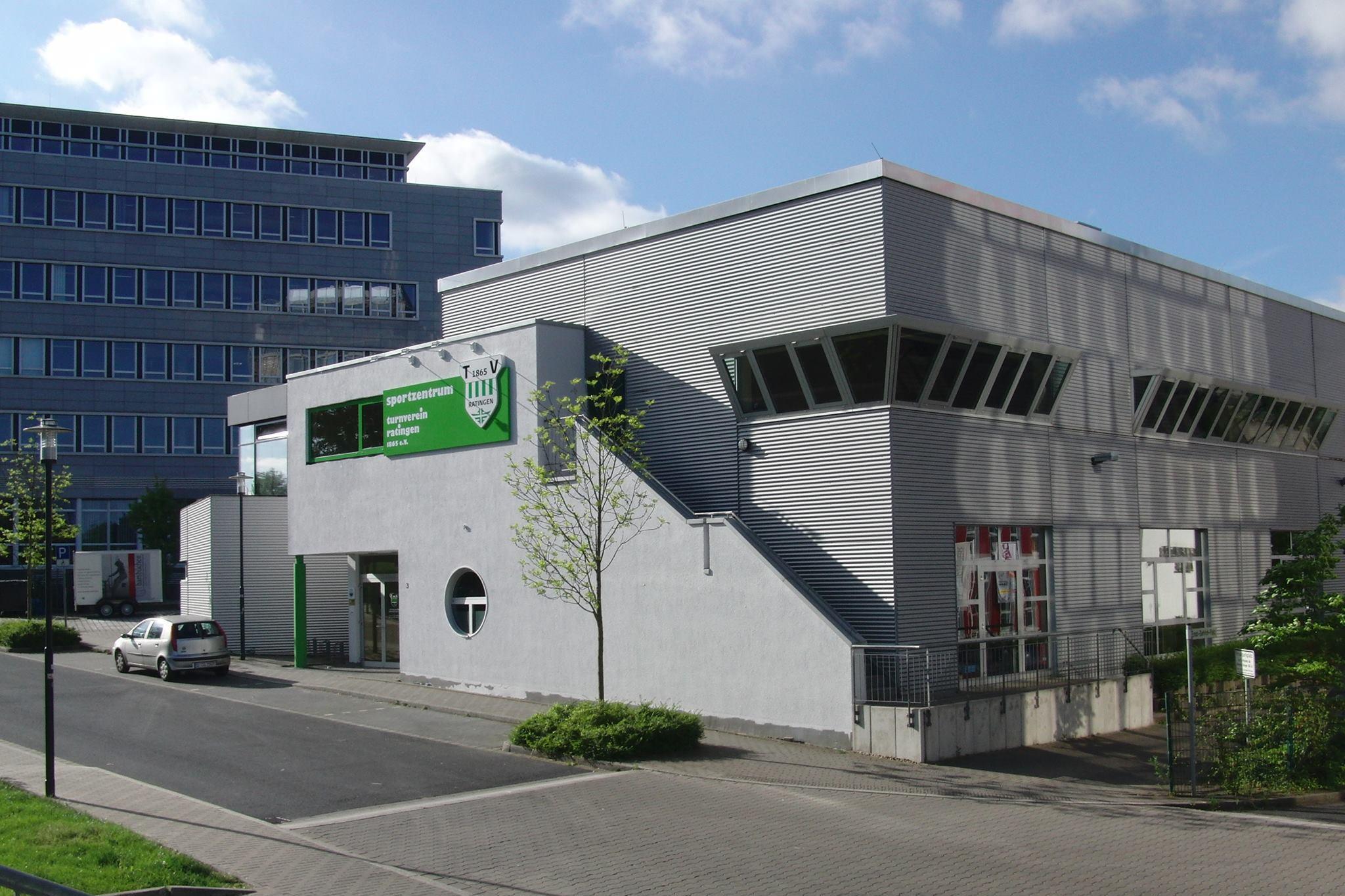 Modernes Sportzentrum mit gr&uuml;ner Beschilderung und grauer Fassade, umgeben von einer gepflasterten Stra&szlig;e und B&auml;umen.