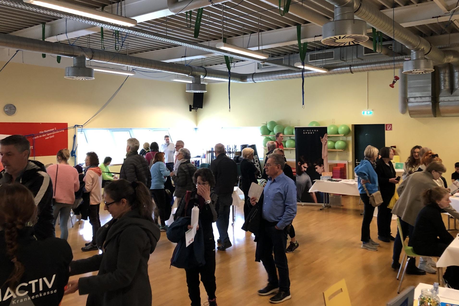 Menschenmenge in einem hellen Raum, der mit Tischen und Info-Ständen für eine Veranstaltung ausgestattet ist.