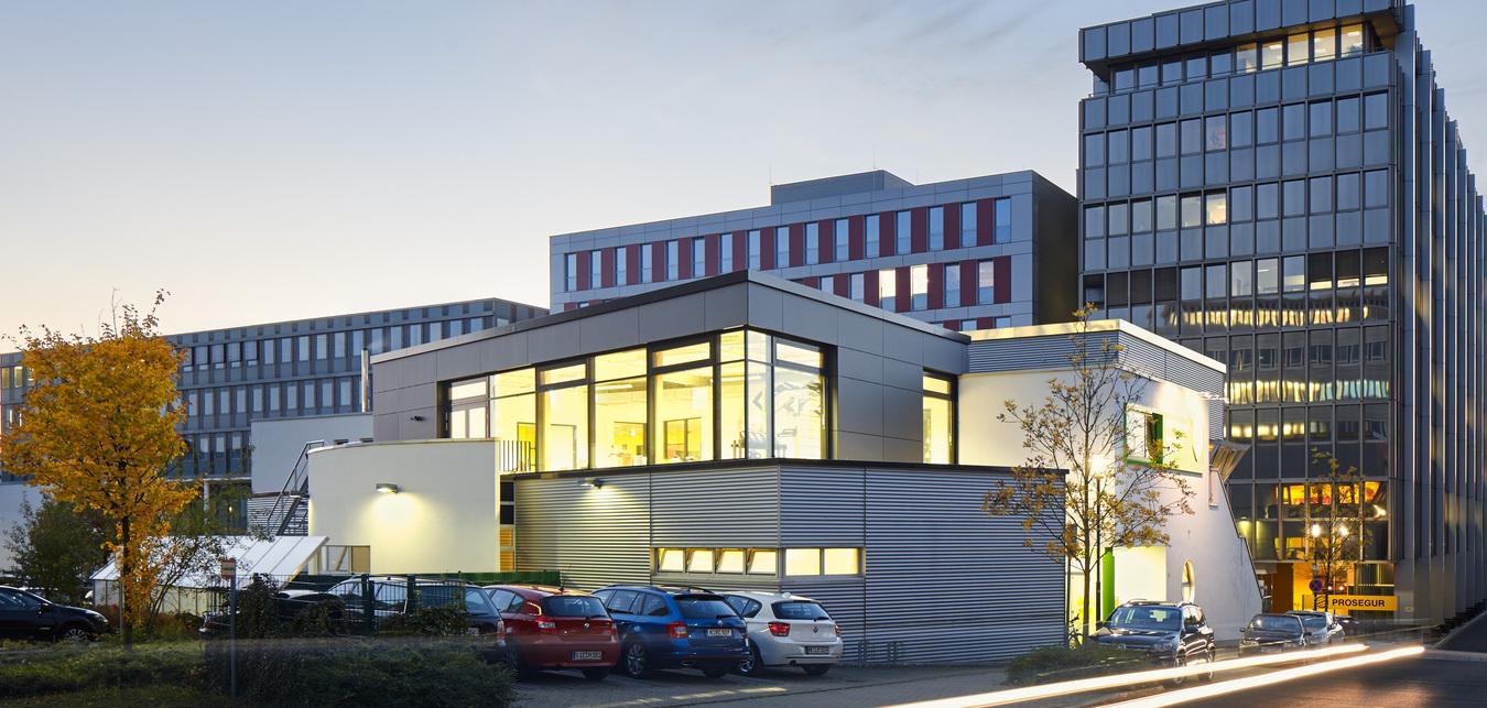 Moderne Geb&auml;ude in Abendd&auml;mmerung, mit beleuchtetem Eintritt und parkenden Autos im Vordergrund.