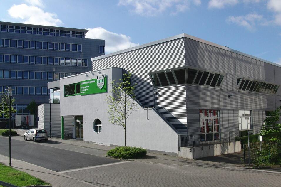 Modernes Sportzentrum mit gr&uuml;ner Beschilderung und grauer Fassade, umgeben von einer gepflasterten Stra&szlig;e und B&auml;umen.