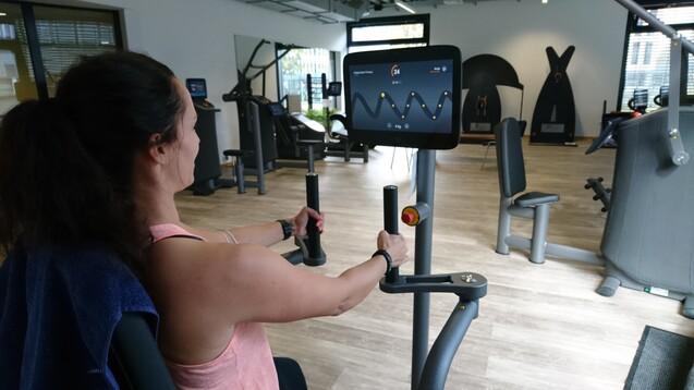 Frau an einem Fitnessger&auml;t mit Bildschirm, w&auml;hrend sie ihr Training in einem modernen Fitnessstudio durchf&uuml;hrt.