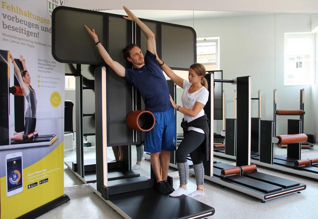 Zwei Personen trainieren mit einem Fitnessger&auml;t in einem modernen Studio, dabei wird eine Dehn&uuml;bung durchgef&uuml;hrt.