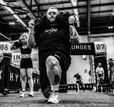 Mann führt einen Ausfallschritt mit Gewichten auf den Schultern in einer Sporthalle aus, während eine Frau hinter ihm trainiert.