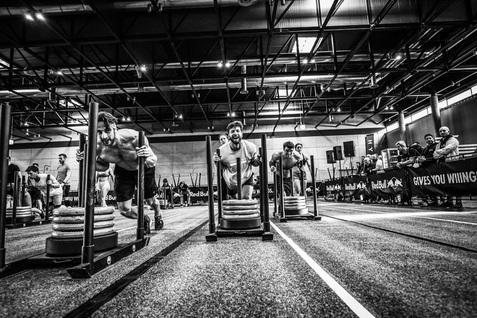 Drei Athleten ziehen Lasten auf Schlitten während eines Wettkampfs in einer großen Halle. Zuschauer beobachten im Hintergrund.