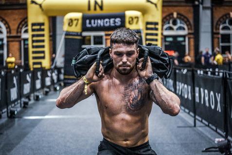 Mann mit tätowiertem Oberkörper trägt eine schwarze Sandsack-Tasche auf den Schultern während eines Wettkampfs.