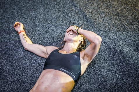 Sportlerin liegend auf Matte, mit einer Hand das Gesicht verdeckend und lacht, in sportlicher Kleidung.
