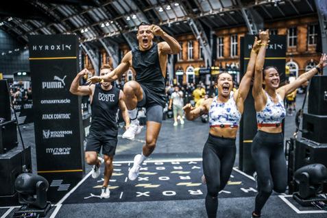 Vier begeisterte Sportler feiern mit Sprüngen und Jubel bei einem Fitness-Event in einer großen Halle.