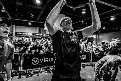 Mann hebt mediales Gewicht über Kopf, während Zuschauer ihn anfeuern, in einem Sportveranstaltungsraum.