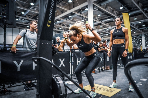 Frau in Sportkleidung trainiert an einer Rudermaschine, beobachtet von anderen Teilnehmern in einem Fitness-Event.