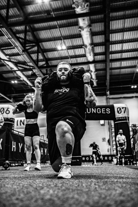 Mann f&uuml;hrt einen Ausfallschritt mit Gewichten auf den Schultern in einer Sporthalle aus, w&auml;hrend eine Frau hinter ihm trainiert.