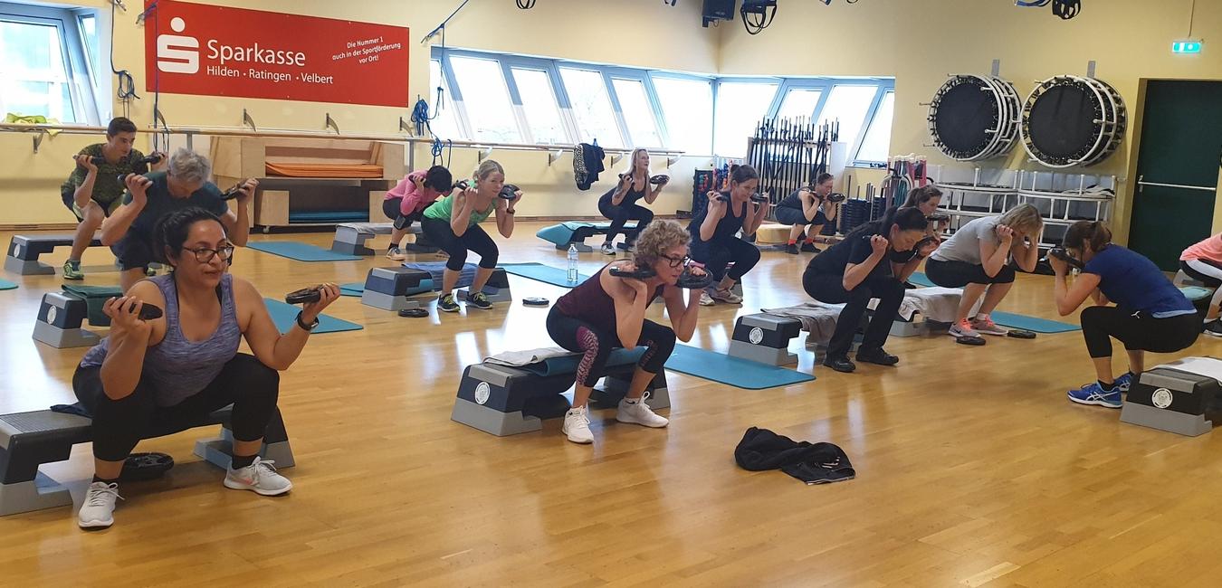 Gruppe von Personen bei einem Fitnesskurs, die mit Gewichten in der Hocke auf Trainingsb&auml;nken arbeiten.