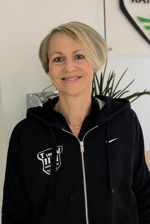 Frau mit kurzem, blonden Haar tr&auml;gt einen schwarzen Rei&szlig;verschluss-Hoodie und steht vor einer Wand mit dem Logo &bdquo;Ratingen&ldquo;.