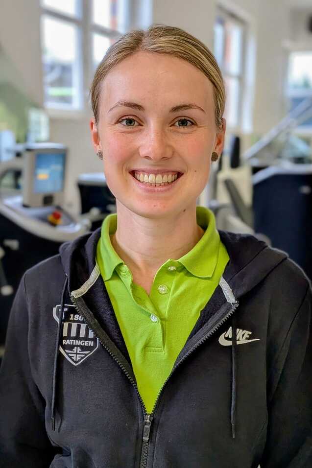 L&auml;chelnde Frau mit kurzen blonden Haaren in Sportkleidung, steht in einem Fitnessstudio mit Trainingsger&auml;ten im Hintergrund.