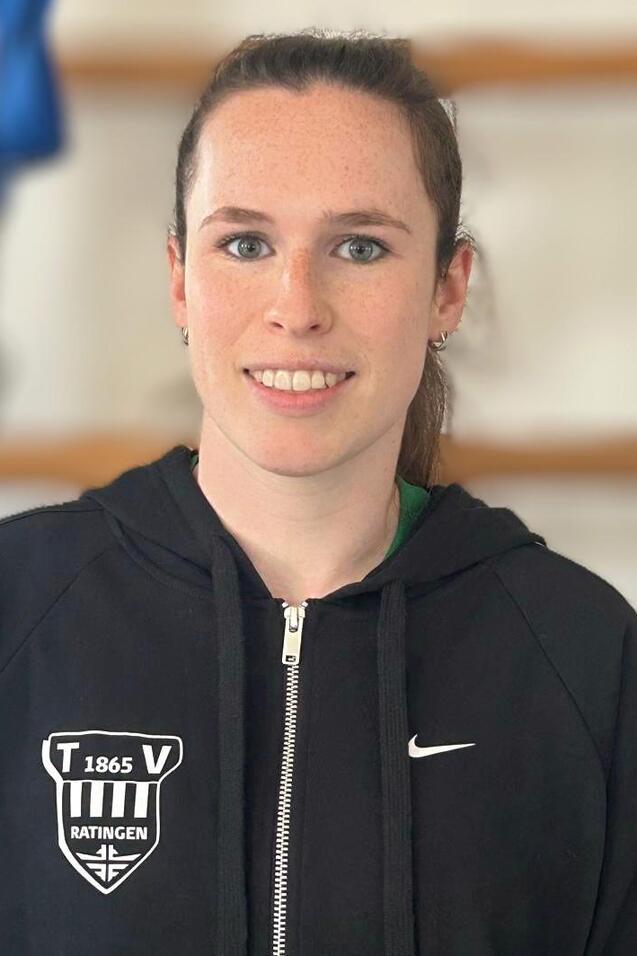Porträt einer sportlichen Frau in schwarzer Jacke mit Logo, lächelnd vor einem neutralen Hintergrund.