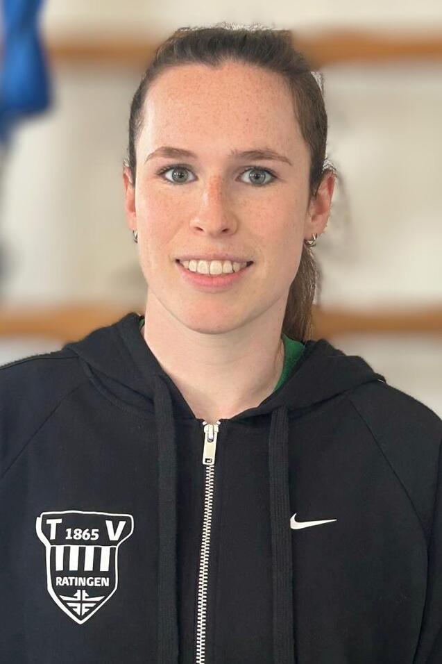 Porträt einer sportlichen Frau in schwarzer Jacke mit Logo, lächelnd vor einem neutralen Hintergrund.