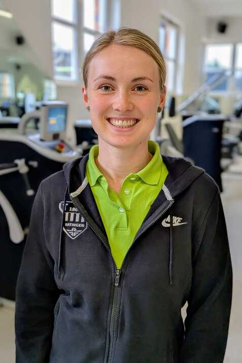 L&auml;chelnde Frau mit kurzen blonden Haaren in Sportkleidung, steht in einem Fitnessstudio mit Trainingsger&auml;ten im Hintergrund.