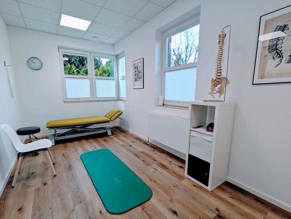 Therapieraum mit gelber Liege, grüner Trainingsmatte, Uhr und anatomischem Skelett auf einem Regal. Helle, freundliche Atmosphäre.