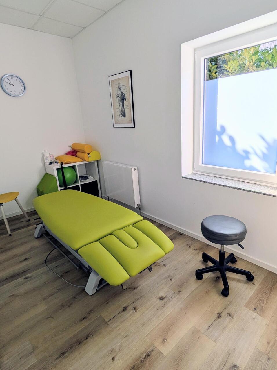 Therapieraum mit grüner Liege, schwarzem Drehstuhl und Fenster, hell erleuchtet durch Tageslicht.